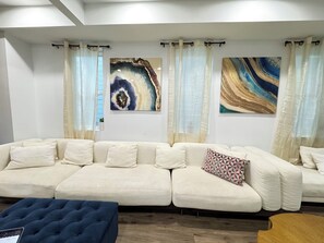 Ein modernes Wohnzimmer mit einem beigefarbenen Ecksofa, zwei abstrakten Gemälden, transparenten Vorhängen und einem dunkelblauen Ottomanen.