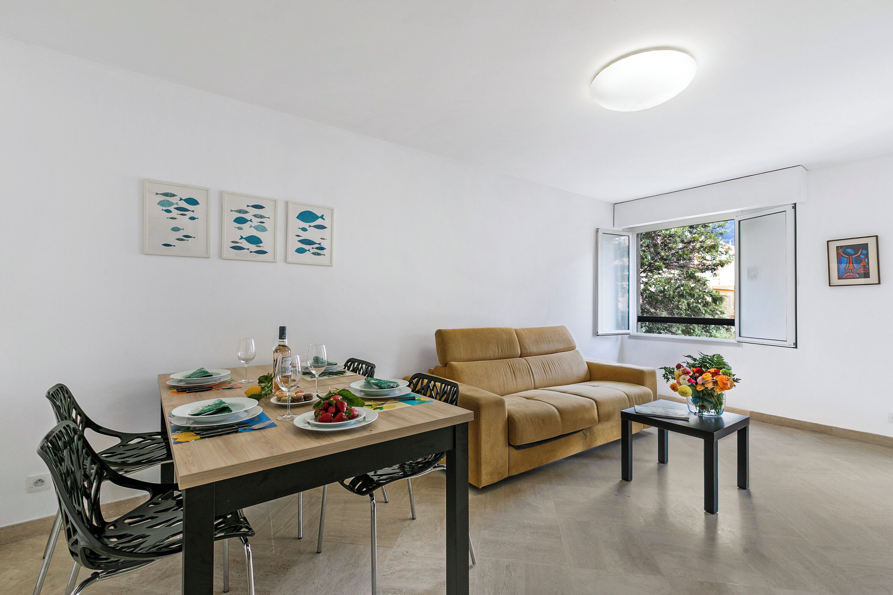 Een eetgedeelte met een tafel voor vier personen, een bank, een klein bijzettafeltje met een vaas, en een raam met uitzicht op groen.