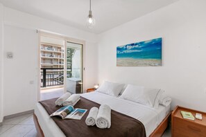 A hotel room with a bed, a nightstand, a reading lamp, a painting of a beach, and a view of a balcony with a railing. - Daturas Bright Apt with Balcony, Menton, France (Menton)
