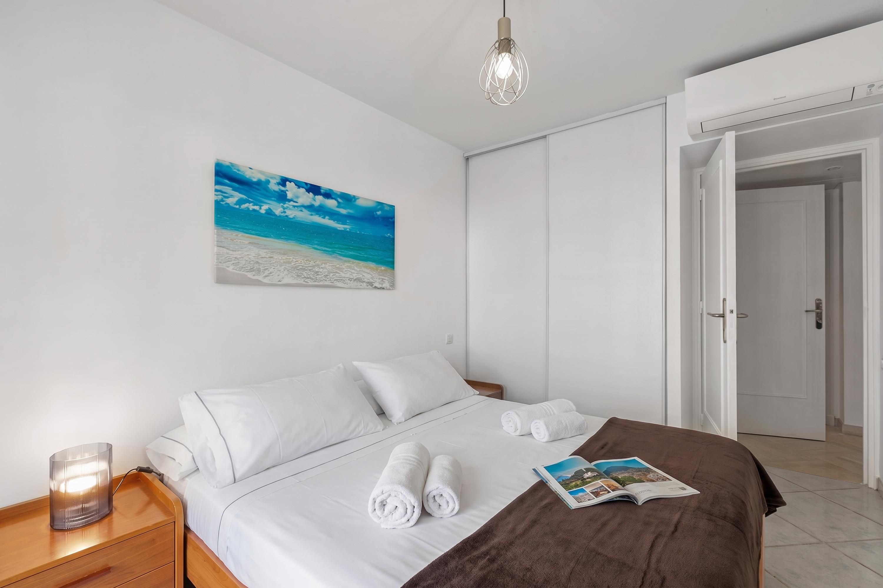 A hotel room with a bed, white pillows, a brown throw blanket, a magazine, and a beach painting on the wall.