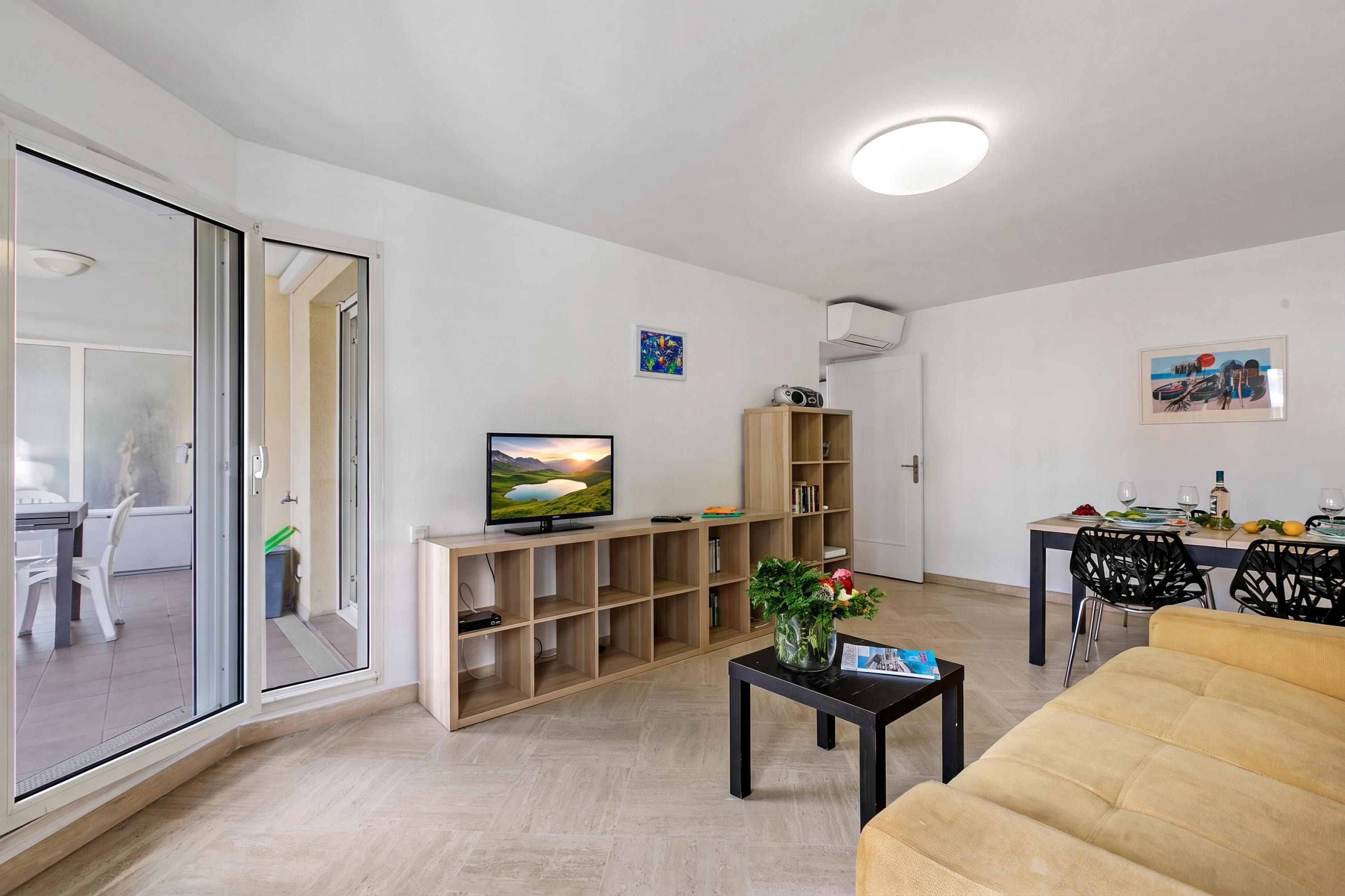 A modern living room with a flat-screen TV, a wooden bookshelf, a dining area with a table and chairs, and a sofa.