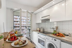 Eine moderne Küche mit Waschmaschine, einem Essbereich mit gedecktem Tisch und Blick auf ein Wohnhaus durch eine offene Tür.