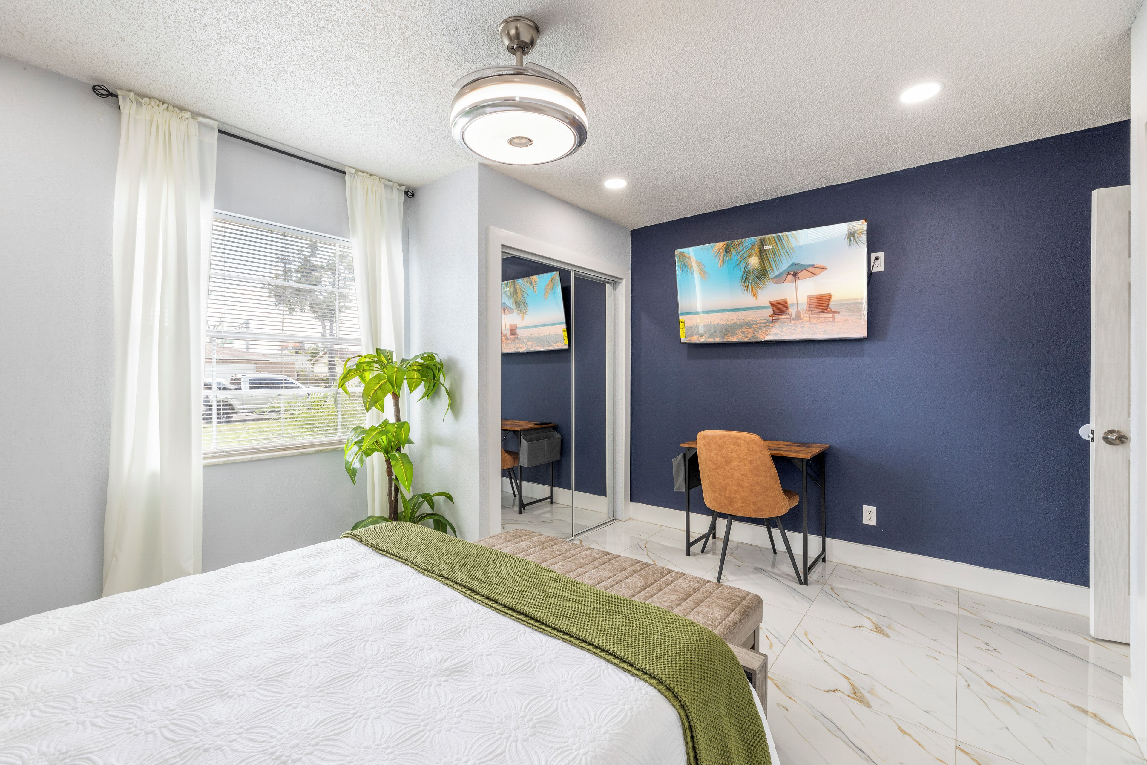 Une chambre d’hôtel avec un lit, un bureau, une chaise, une plante en pot et un tableau au mur.