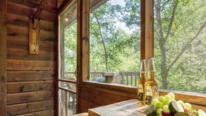 Interior de una cabaña de madera con una mesa de madera, una ventana con vista a árboles y botellas de alcohol.