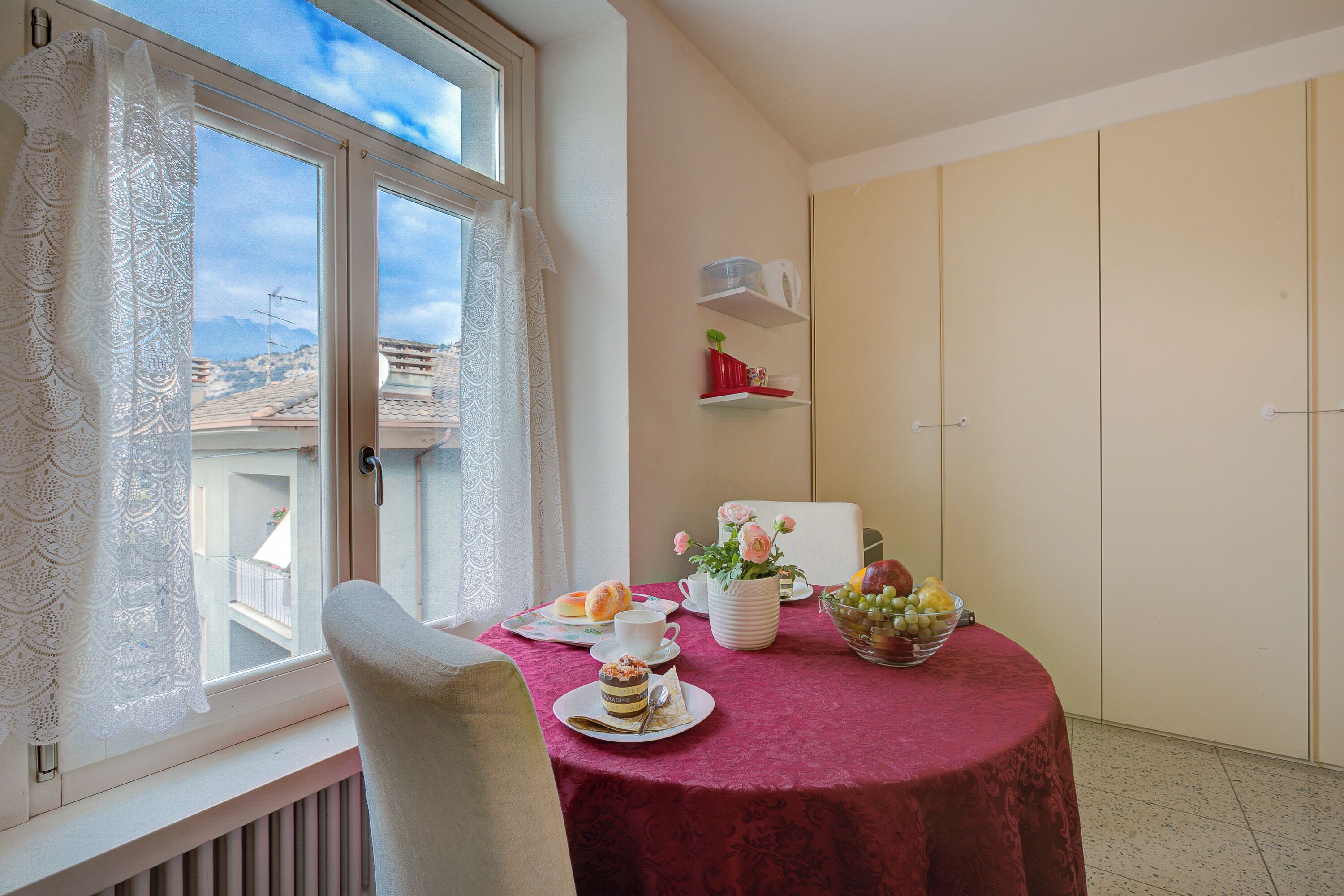 A dining area with a table set for two, a window with lace curtains, a view of the city, and a wardrobe.