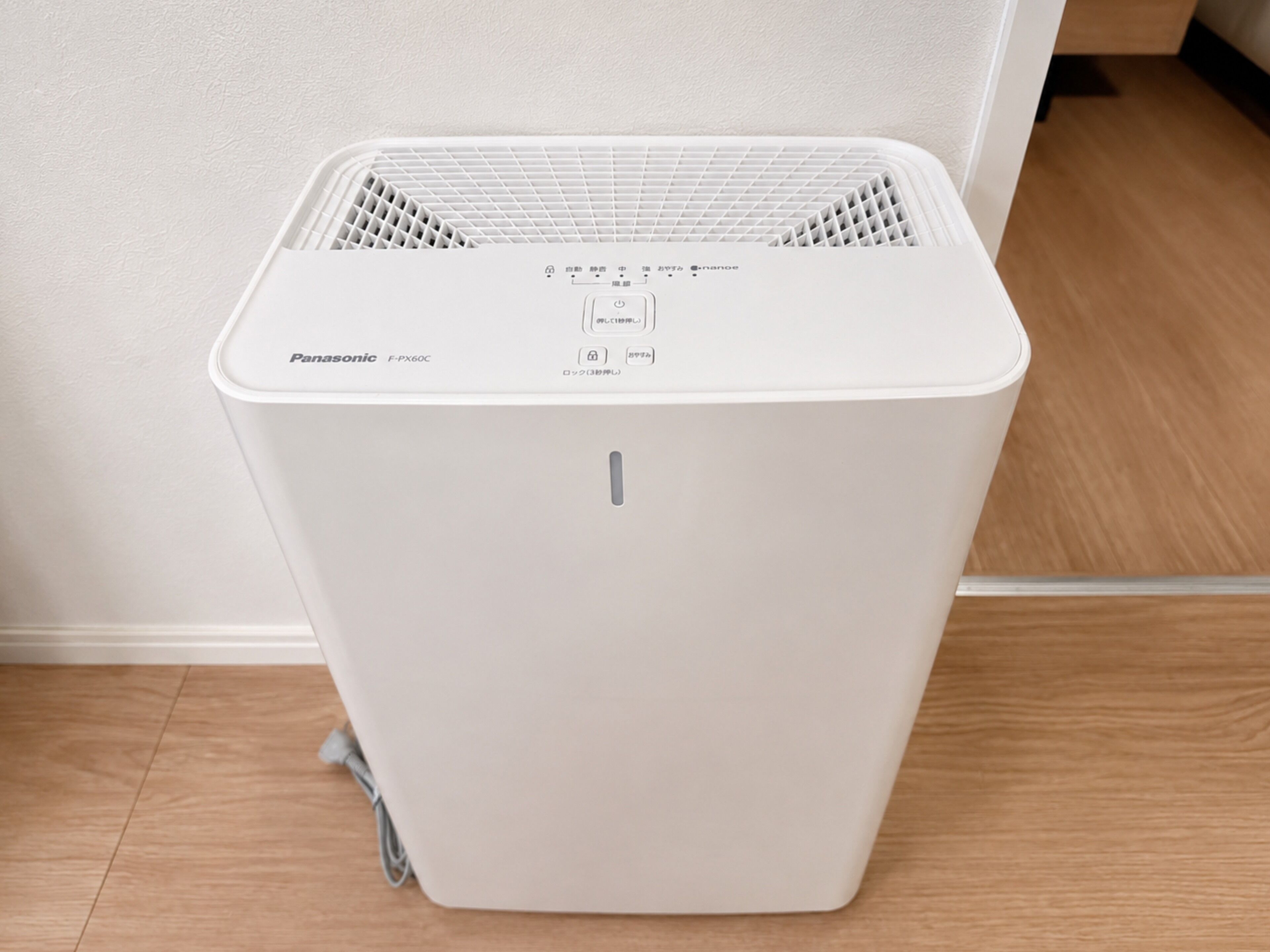 A white Panasonic F-PX60C portable air purifier with a control panel, placed on a wooden floor against a white wall.