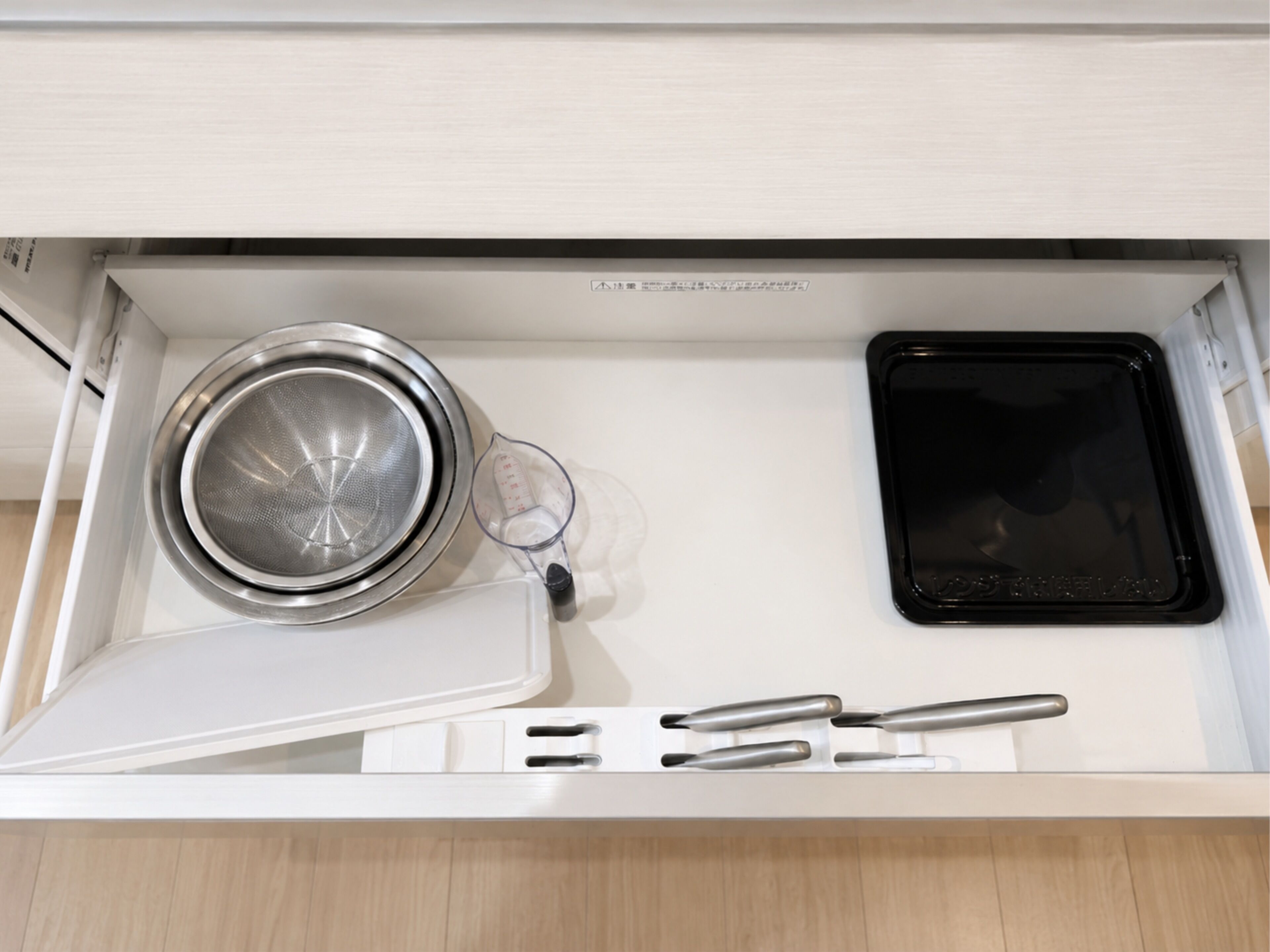 A kitchen drawer with a stainless steel strainer, a black microwave, and a measuring cup.