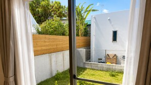Visão através de uma janela com cortinas, revelando uma área externa gramada com cerca de madeira, uma parede de concreto e uma pequena estrutura com janela.