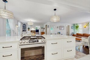 Eine geräumige Küche mit einer großen Kochinsel, weißen Schränken und einem eingebauten Backofen. Über der Insel hängen Pendelleuchten und es gibt einen Essbereich mit einem braunen Sofa.