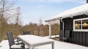 A covered outdoor seating area with a table and chairs, a window with yellow lamps, and a snow-covered deck. - 4 Star Holiday Home in Knebel (Knebel)