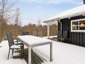 Ein überdachter Außenbereich mit Tisch und Stühlen, ein Fenster mit gelben Lampen und eine mit Schnee bedeckte Terrasse.