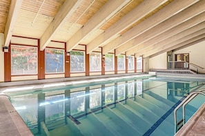 An indoor swimming pool with wooden beams, large windows, and a staircase leading to an upper level.