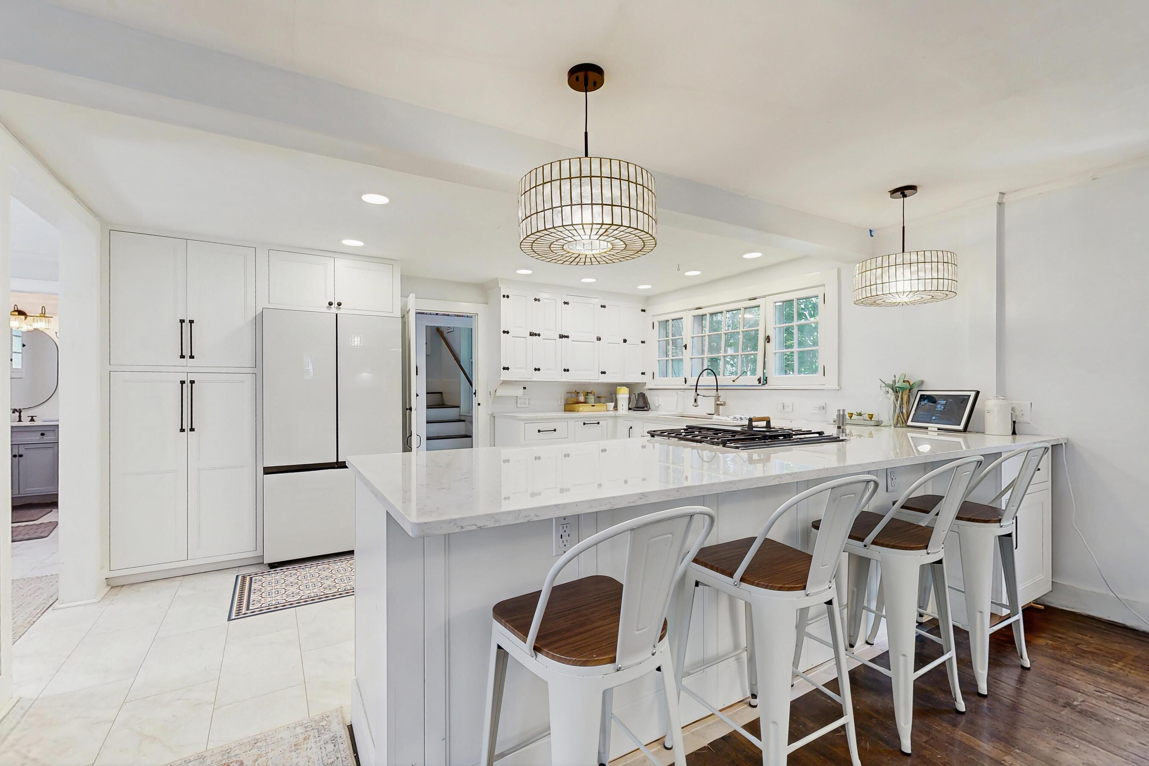 A modern kitchen with white cabinetry, a central island with bar stools, pendant lighting, and a view to the outside.