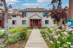 A two-story building with a front porch, a wooden pathway leading to the entrance, and a well-maintained garden with pink flowers. - Mt. Snow All Season Condo: w/ shared hot tub & pool, ski shuttle, tennis (Dover)