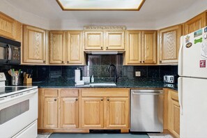 A kitchen with wooden cabinets, a black countertop, a stainless steel sink, and a refrigerator with magnets. - Condo with gas fireplace and hiking trails at the door, near Story Land (Bartlett)