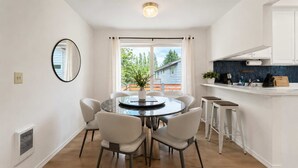 Et moderne køkken med hvide skabe, et rundt spisebord med stole, et stort vindue med gardiner og en lille ø med barstole.