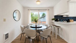 Eine moderne Küche mit weißen Schränken, einem runden Esstisch mit Stühlen, einem großen Fenster mit Vorhängen und einer kleinen Kochinsel mit Hockern.