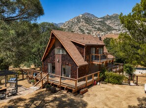 Una cabaña con terraza de madera, balcón y montañas al fondo.