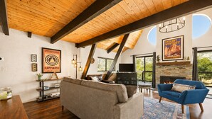 Een ruime woonkamer met een hoge plafond, houten balken, een open haard en een groot raam met uitzicht op groen.