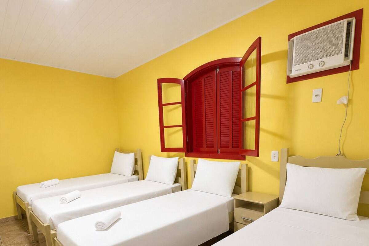 Ein Hotelzimmer mit drei Einzelbetten, einem roten Fensterladen, einer Klimaanlage und einem gefliesten Boden.