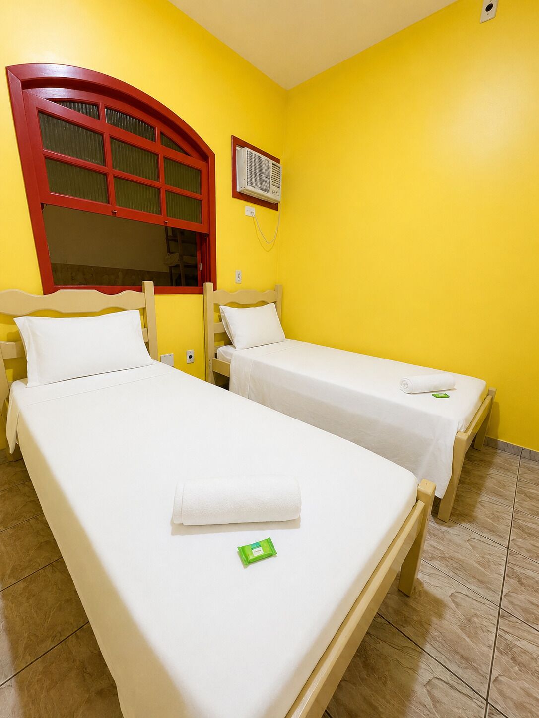 A room with two single beds, a red-framed window, an air conditioning unit, and a tiled floor.