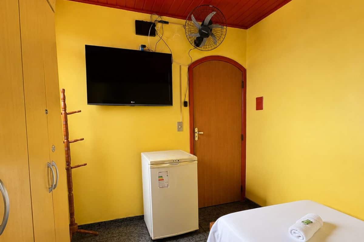 Ein Zimmer mit einem Bett, einem Fernseher, einem Ventilator, einer Tür und einem kleinen Kühlschrank.