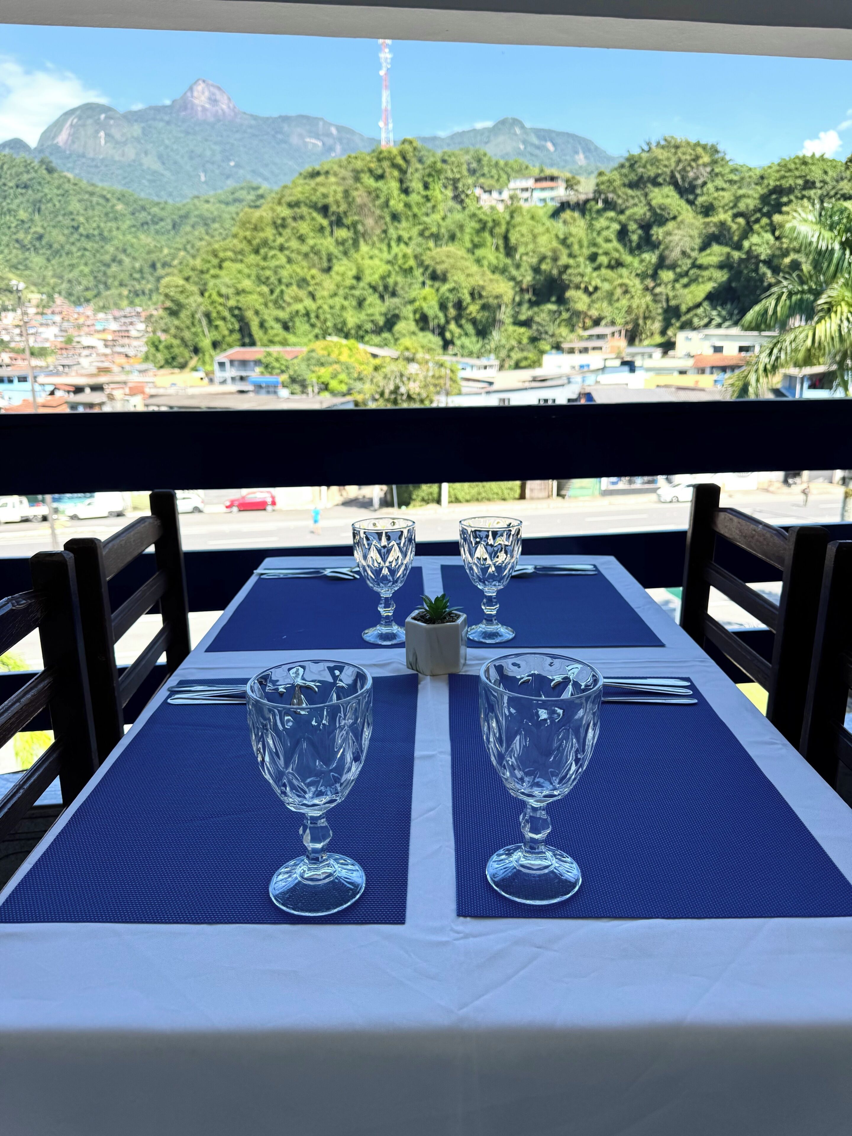 Mesa servida con cuatro copas de vino y una planta pequeña, con vistas a un paisaje montañoso y edificios a lo lejos.