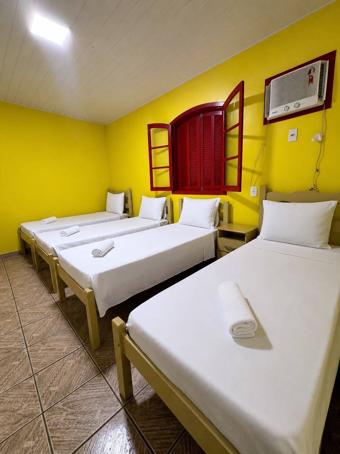 A hotel room with three single beds, a red-framed window, and a wall-mounted air conditioning unit.