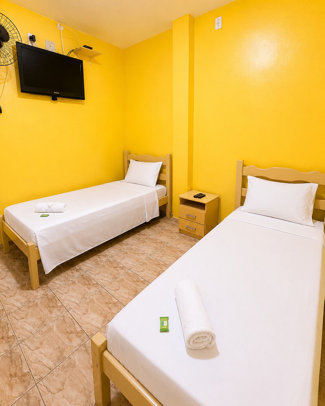 Habitación de hotel con dos camas individuales, un televisor montado en la pared, una mesita de noche pequeña y suelo de baldosas.
