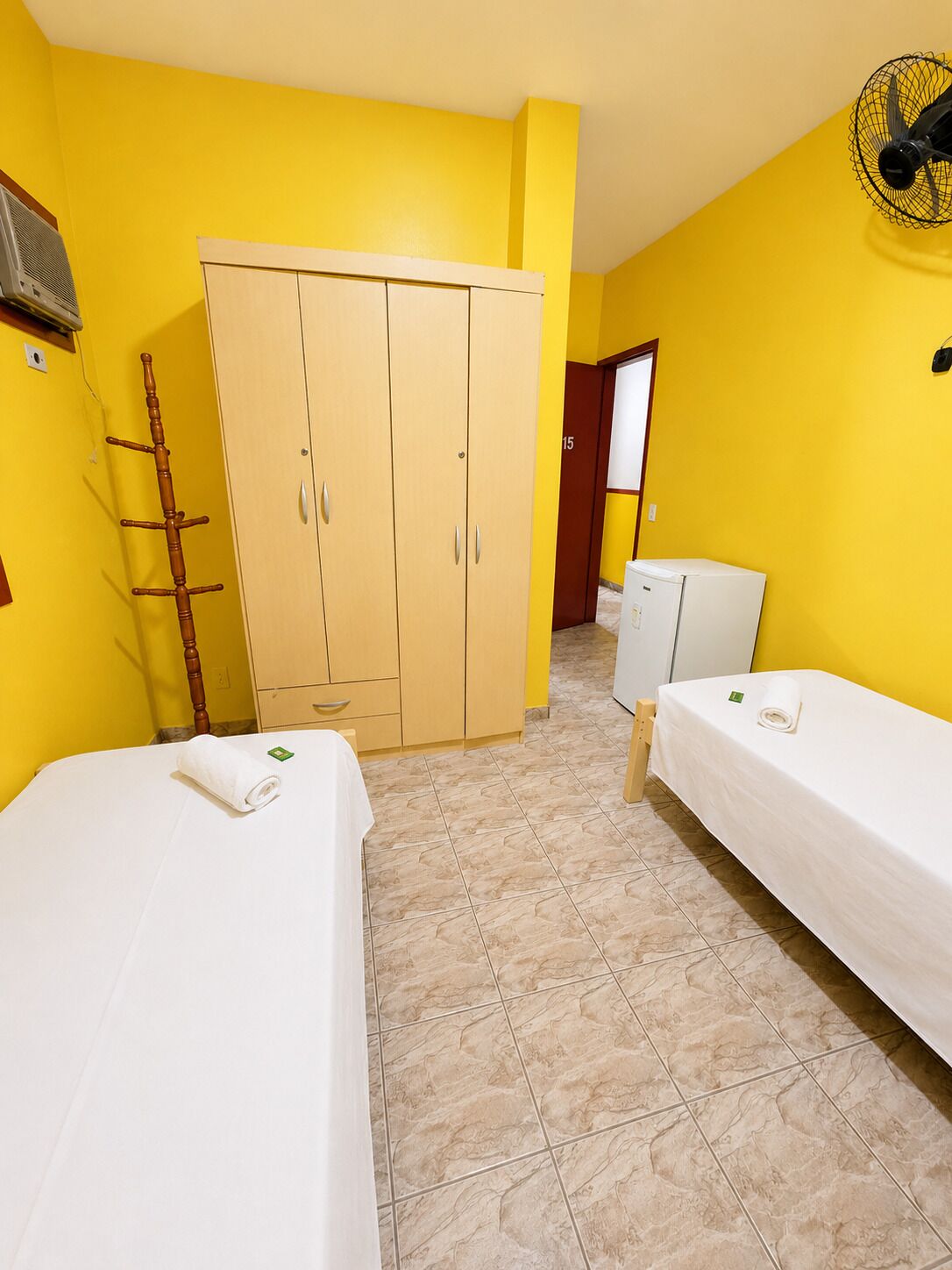 A room with two beds, a wardrobe, a fan, and a small refrigerator.