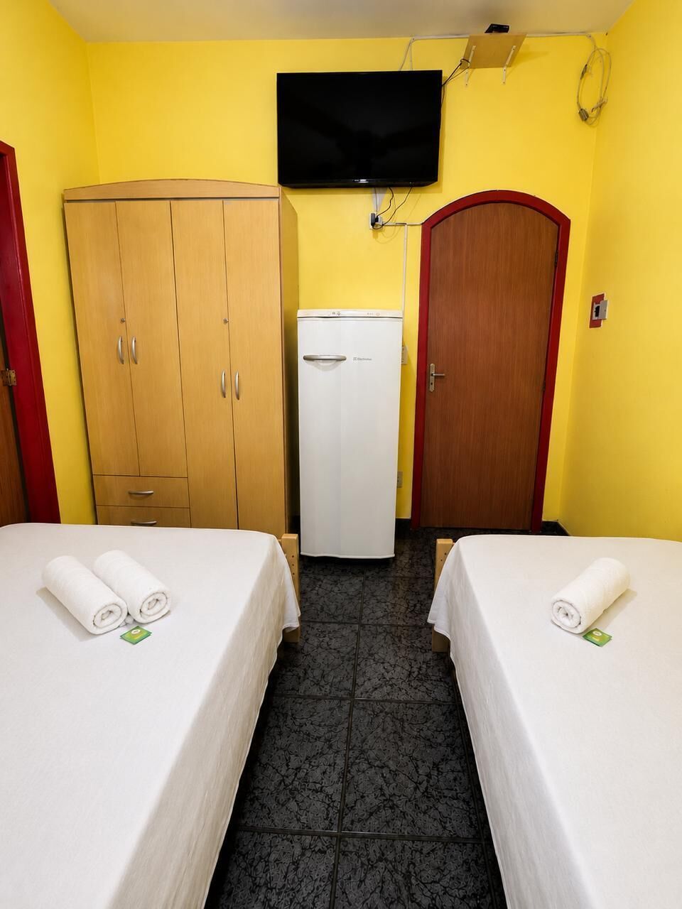 Hotellihuone, jossa on kaksi sänkyä, vaatekaappi, jääkaappi ja seinälle asennettu televisio.