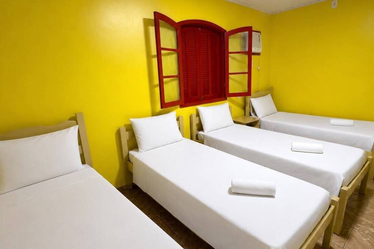 Ein Hotelzimmer mit drei Einzelbetten, jedes mit weißer Bettwäsche und einem aufgerollten Handtuch. Die Wände sind gelb, und über den Betten befindet sich ein Fenster mit rotem Rahmen.