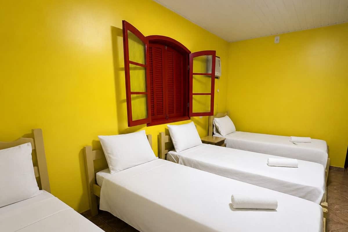 Ein Hotelzimmer mit vier Einzelbetten, einem fenster mit rotem Rahmen und einer weißen Klimaanlage an der Wand.