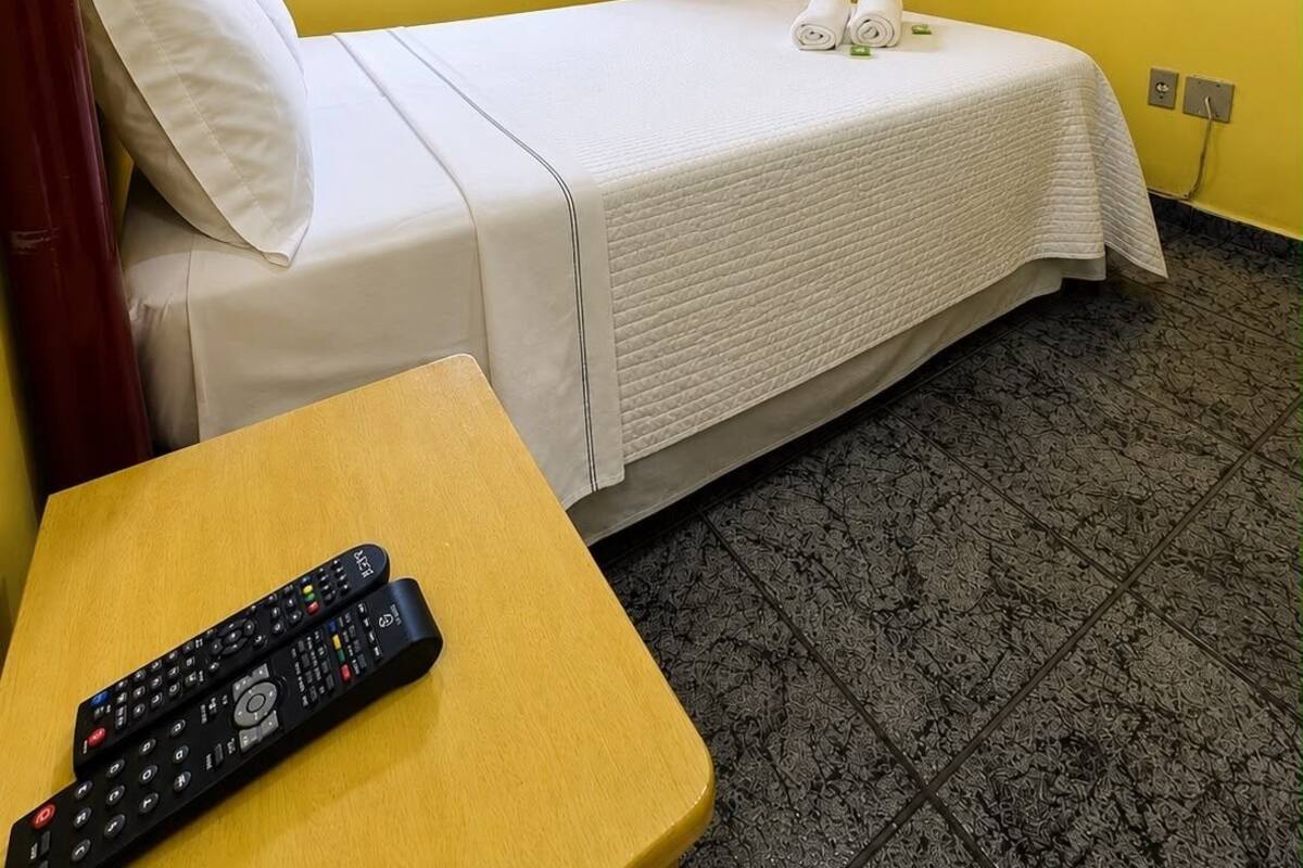 Ein Einzelbett mit weißer Bettdecke, ein an der Wand befestigter Fernseher, ein Fenster mit roten Rollläden und eine Fernbedienung auf einem Holztisch.