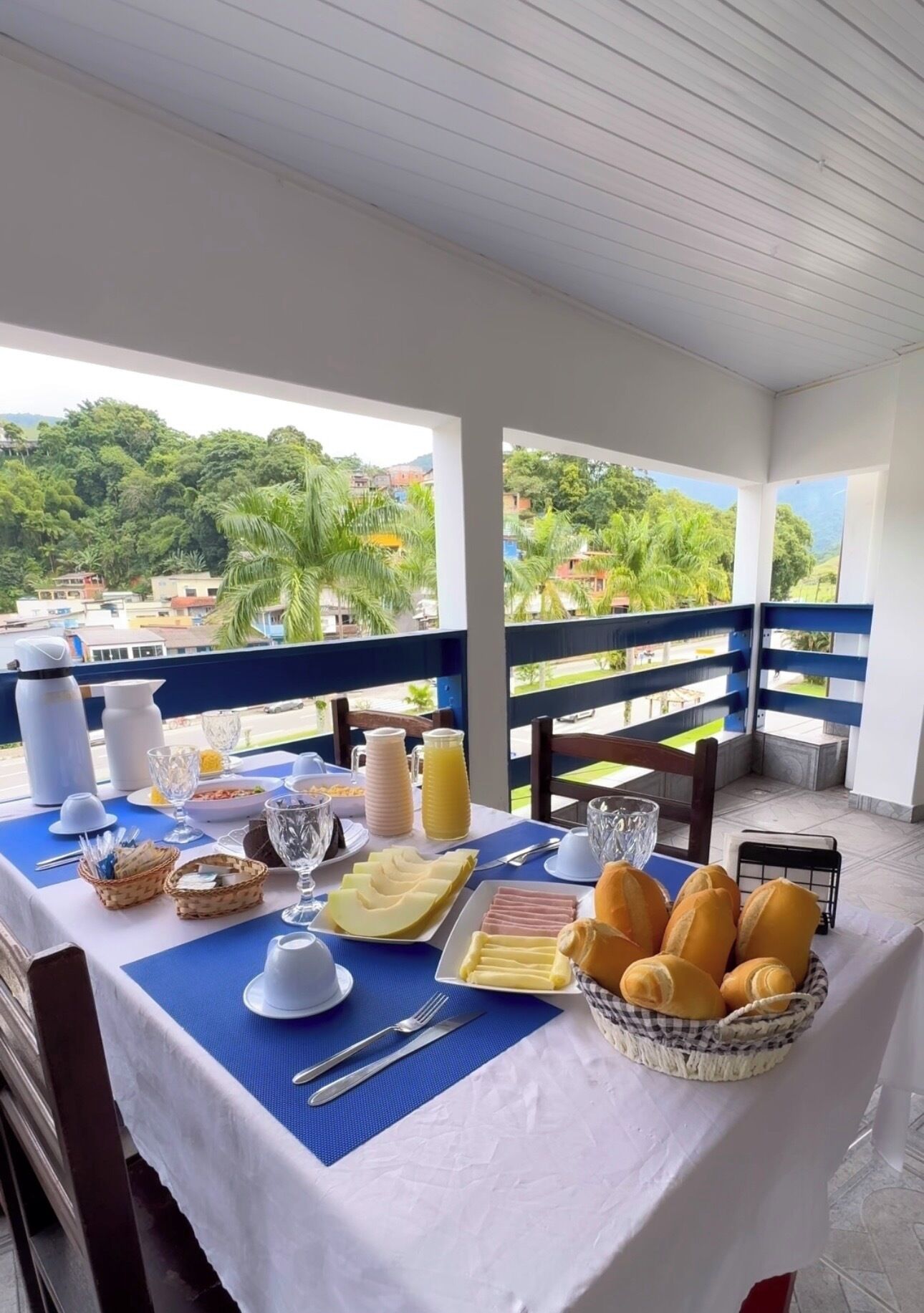 Una mesa preparada con alimentos para el desayuno, vista a un paisaje verde y barandilla de balcón.