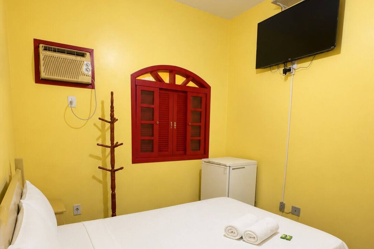 Ein Einzelbett mit weißer Bettwäsche, ein Fenster mit rotem Rahmen, eine weiße Heizung und ein Flachbildfernseher an der Wand.