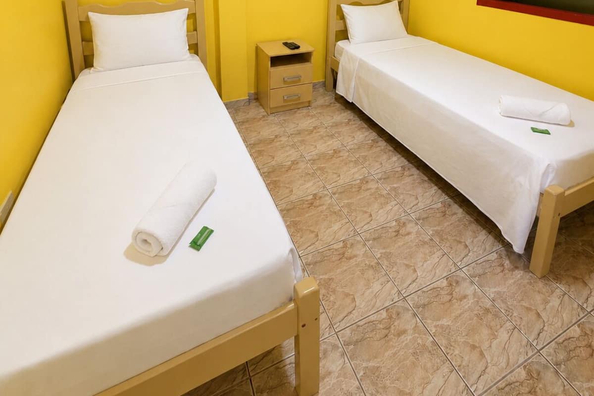 Ein Zimmer mit zwei Einzelbetten, einem kleinen Nachttisch, einem Beistelltisch und einem Fenster mit rotem Rahmen.