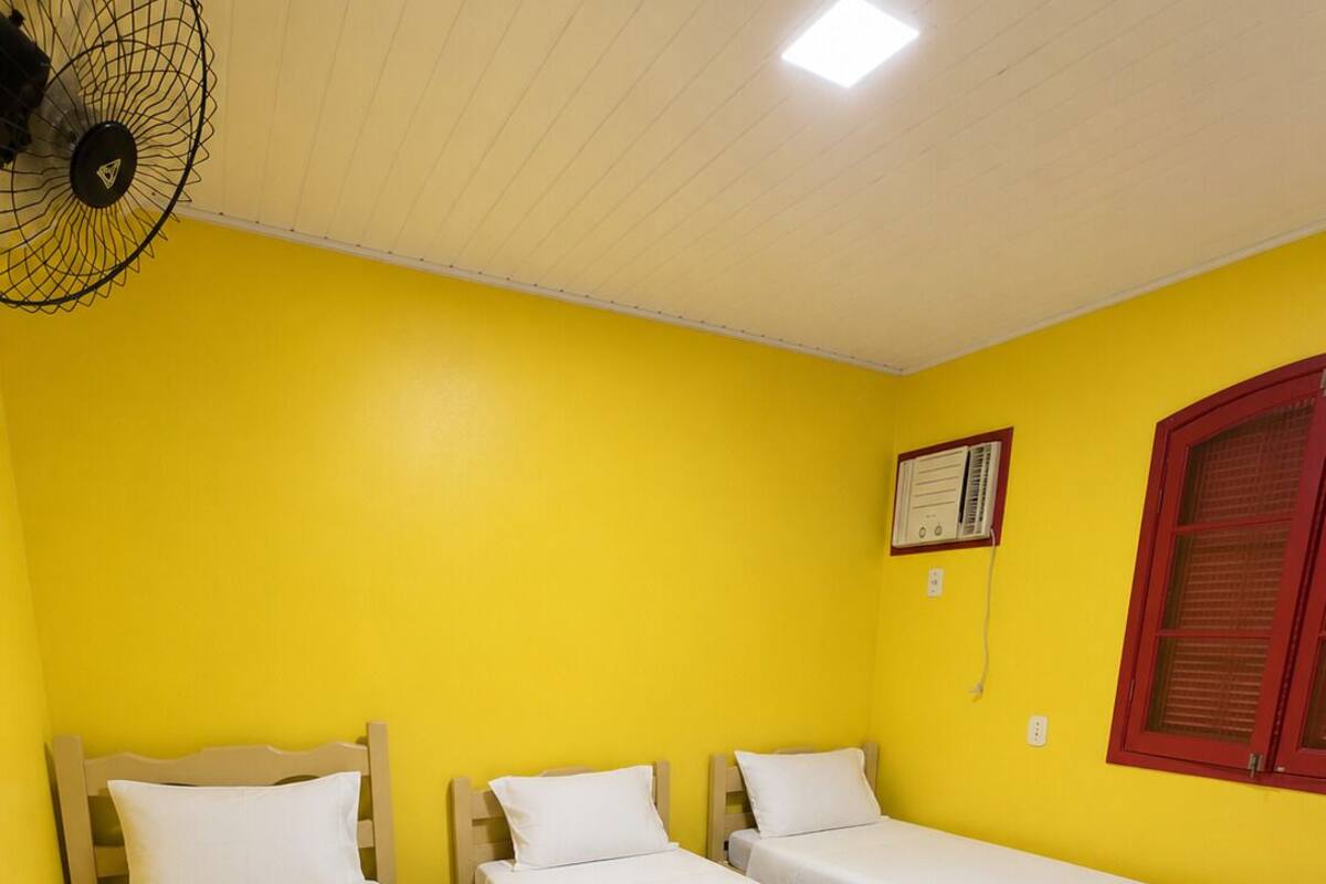 Ein Zimmer mit drei Einzelbetten, einem Deckenventilator, einem Heizkörper und einem Fenster mit Jalousien.