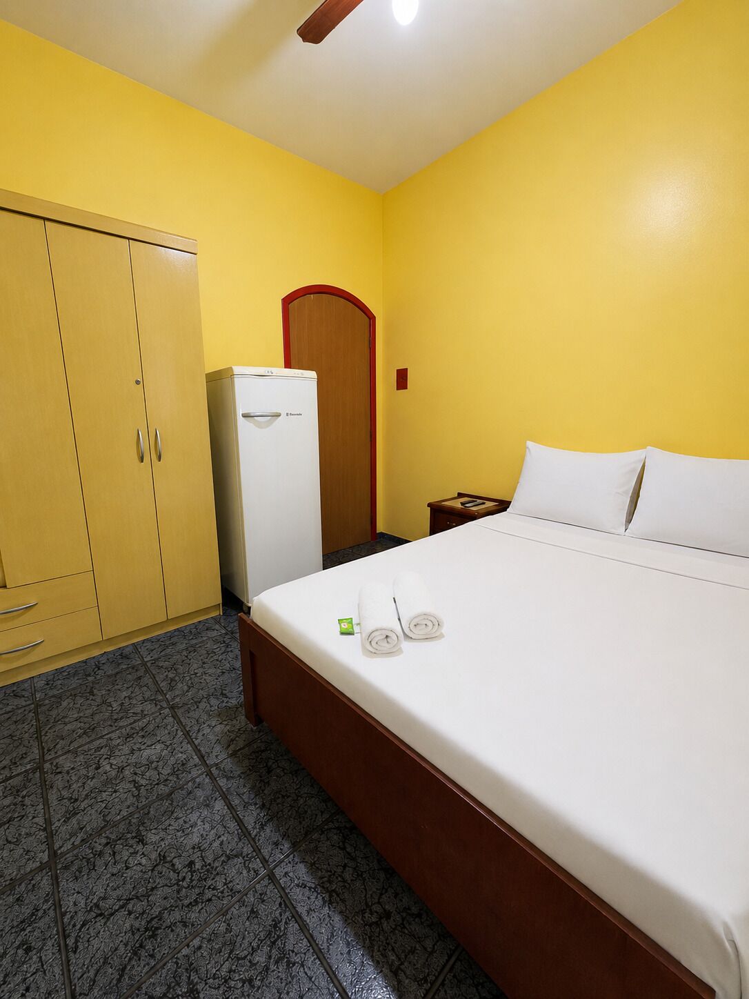Una habitación de hotel con una cama, un armario, un refrigerador y una puerta de madera.