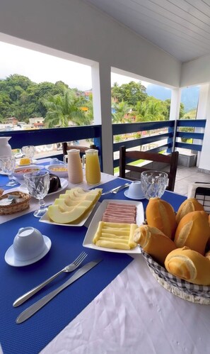 A breakfast table with food and drinks, a view of greenery and buildings, and a balcony railing.