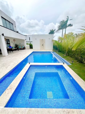 A rectangular swimming pool with a tiled edge, a white wall with a door, and a seating area with a sofa and table. - Luxury Villa in Punta Cana - Casa Monika (Punta Cana)