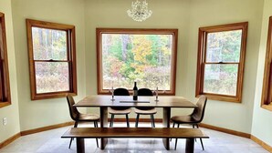 A dining area with a wooden table, four chairs, a bench, and three windows with autumn foliage views. - Fire Pit Evenings • Outdoor Dining - Five Acres (Palenville)