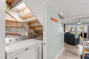 A laundry area with a washing machine, dryer, and a stove. There is a ceiling fan, a window with a view, and a chair in the adjacent living area. - Steps to Beach, Pool, Pickleball & 3BR Sleeps 6 (Panama City Beach)