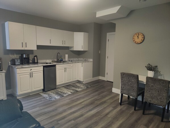 A modern kitchen with white cabinets, a built-in dishwasher, and a coffee maker. There's a dining area with a table and chairs, a clock on the wall, and a potted plant.