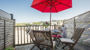 Um deck de madeira com um guarda-sol vermelho, duas cadeiras de madeira, uma mesa com um vaso de flores e vista para casas ao fundo.