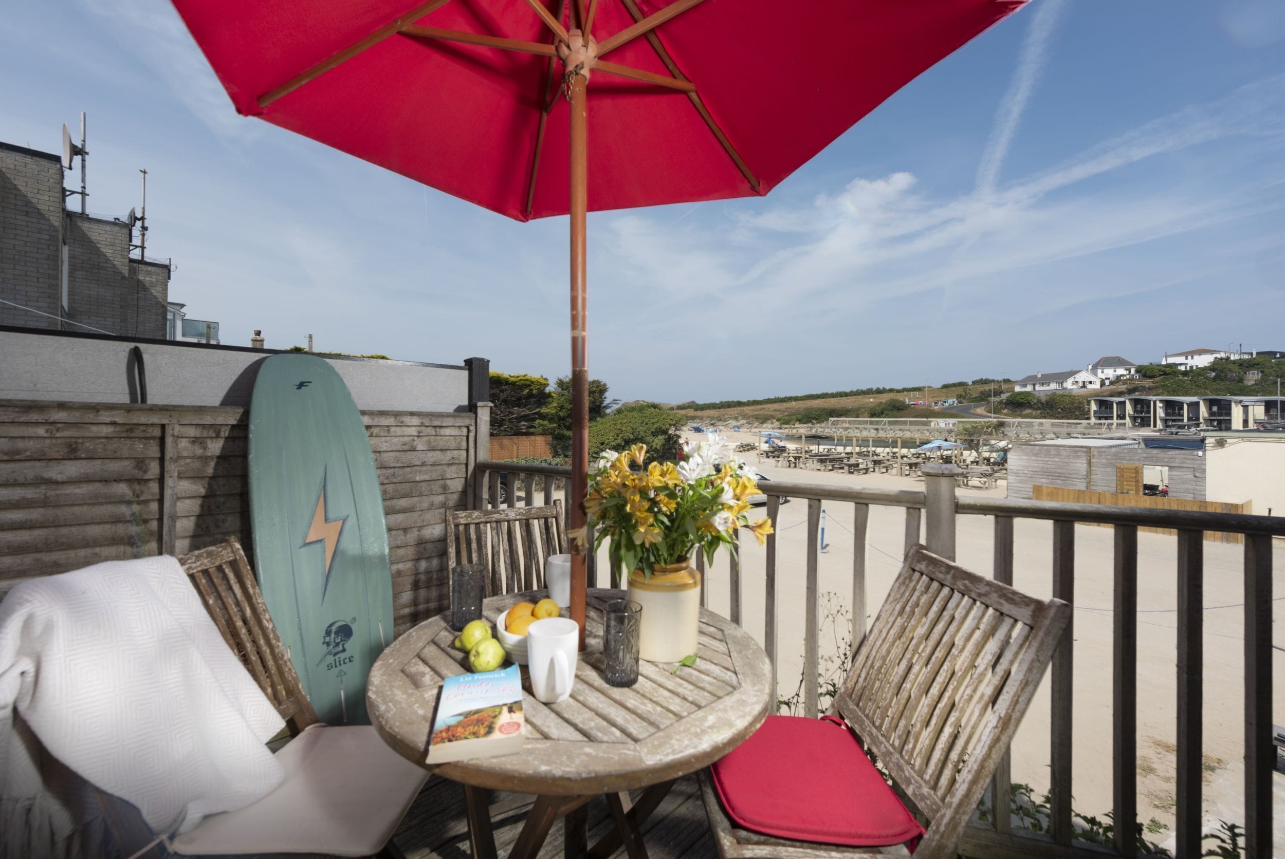 Ein Balkon mit einem roten Sonnenschirm, einem Surfbrett, einem Tisch mit einem Buch und Blick auf den Strand.