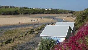 Vista costeira com praia, um pequeno edifício com telhado branco e vegetação ao fundo.