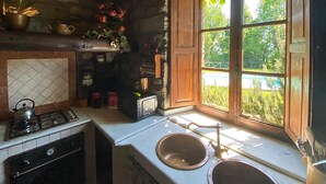 Una cocina rústica con pared de piedra, armarios de madera, estufa a gas, lavamanos y ventana con vistas a follaje.
