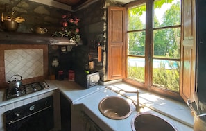 A rustic kitchen with a stone wall, wooden cabinets, a gas stove, a sink, and a window overlooking greenery. - Lovely home in Villa Collemandina (Villa Collemandina)