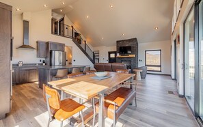 Un vaste espace de vie comprenant une grande table à manger en bois, des sièges confortables, une cheminée et une vue dégagée grâce à de grandes fenêtres.